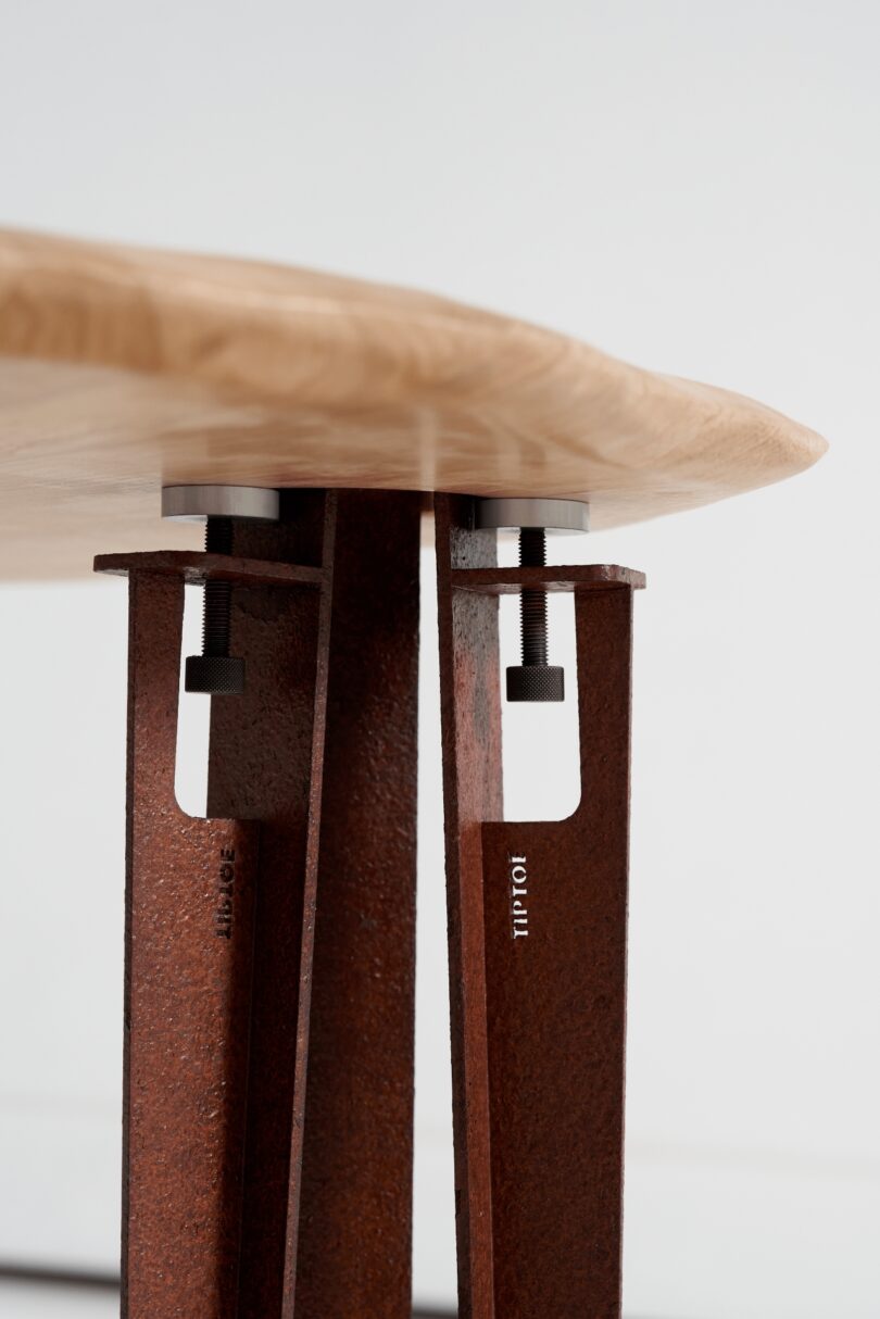 Close-up view of a wooden table top supported by metal legs with visible bolts and rust texture, as if balanced on tiptoe for a subtle industrial charm