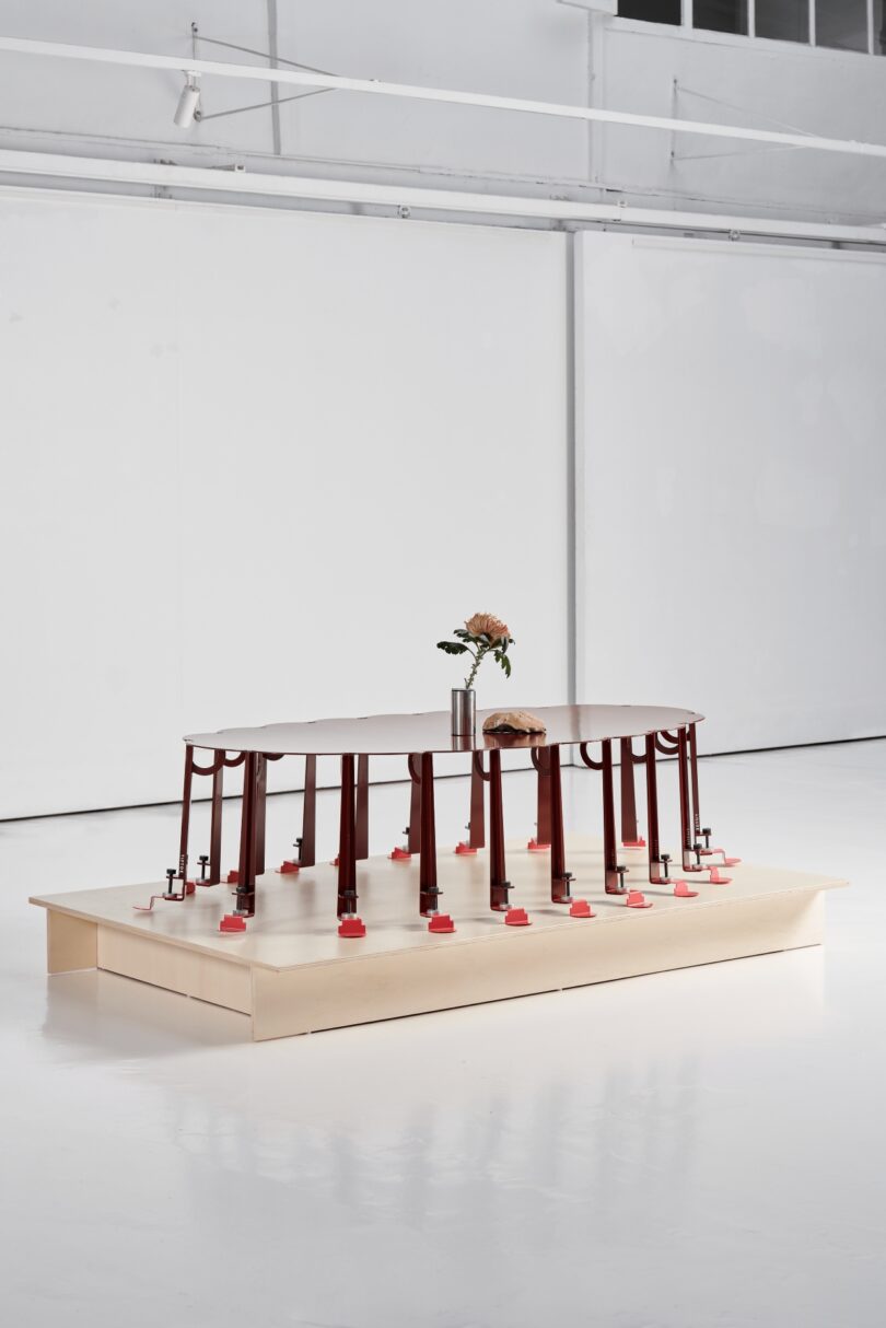 A table supported by red-legged clamps stands tiptoe on a wooden platform in a white room, with a metal vase holding flowers and a loaf of bread resting on its surface