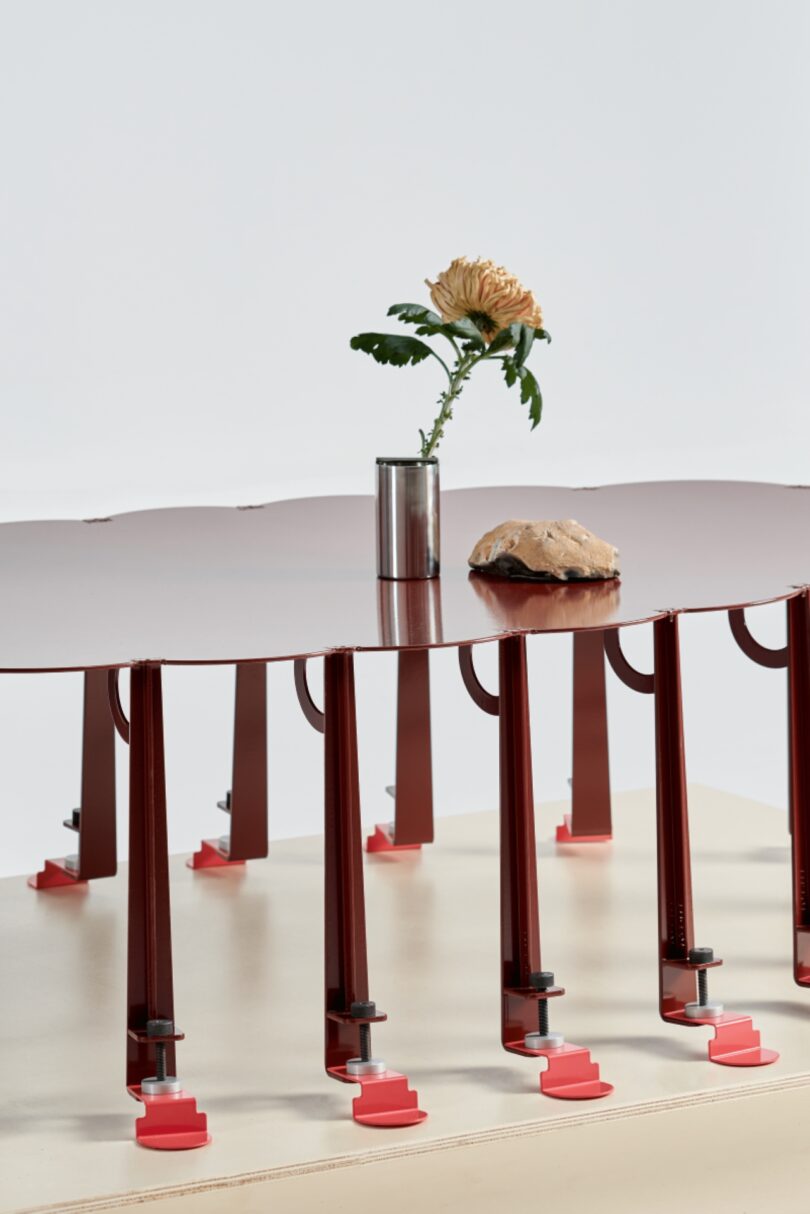 A red table with multiple legs stands ready, as if on tiptoe, with a single flower in a metal vase and a rock resting on top