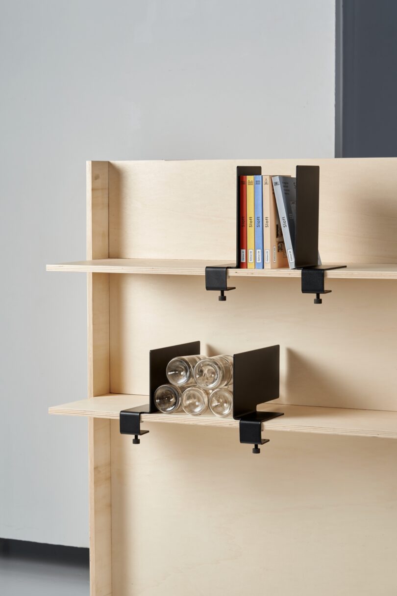 Two wooden shelves with black metal bookends hold several books and four glass cups stacked tiptoe-like. The background is plain and gray