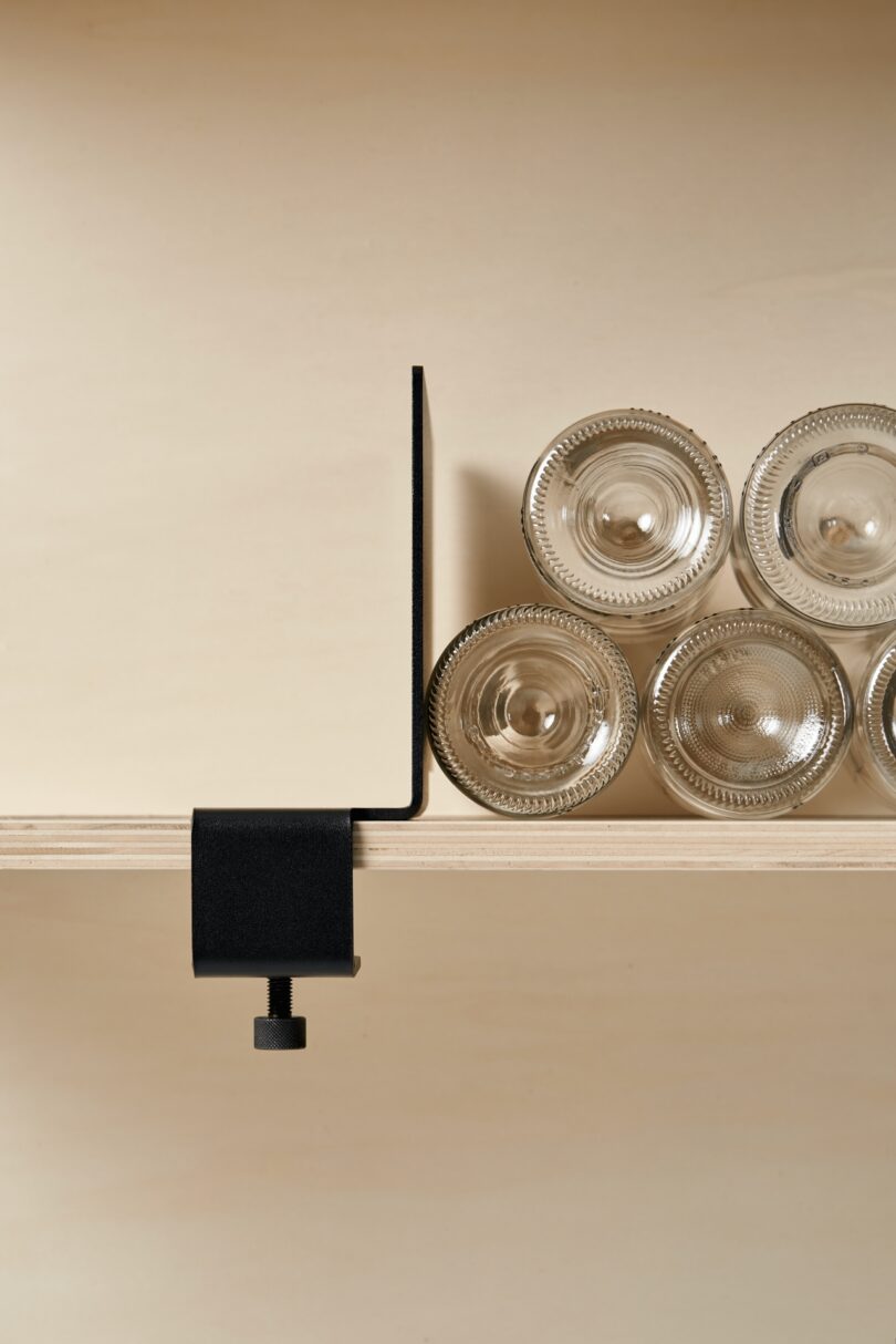 A black metal clamp tiptoes along the edge of a wooden shelf next to five upside-down clear glass cups