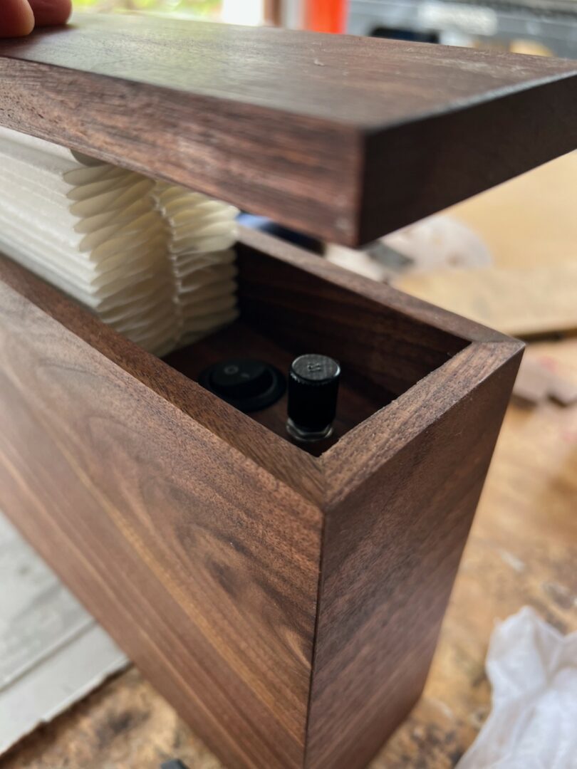 A close-up of a wooden box with a hinged lid, holding folded white paper and two small switches inside