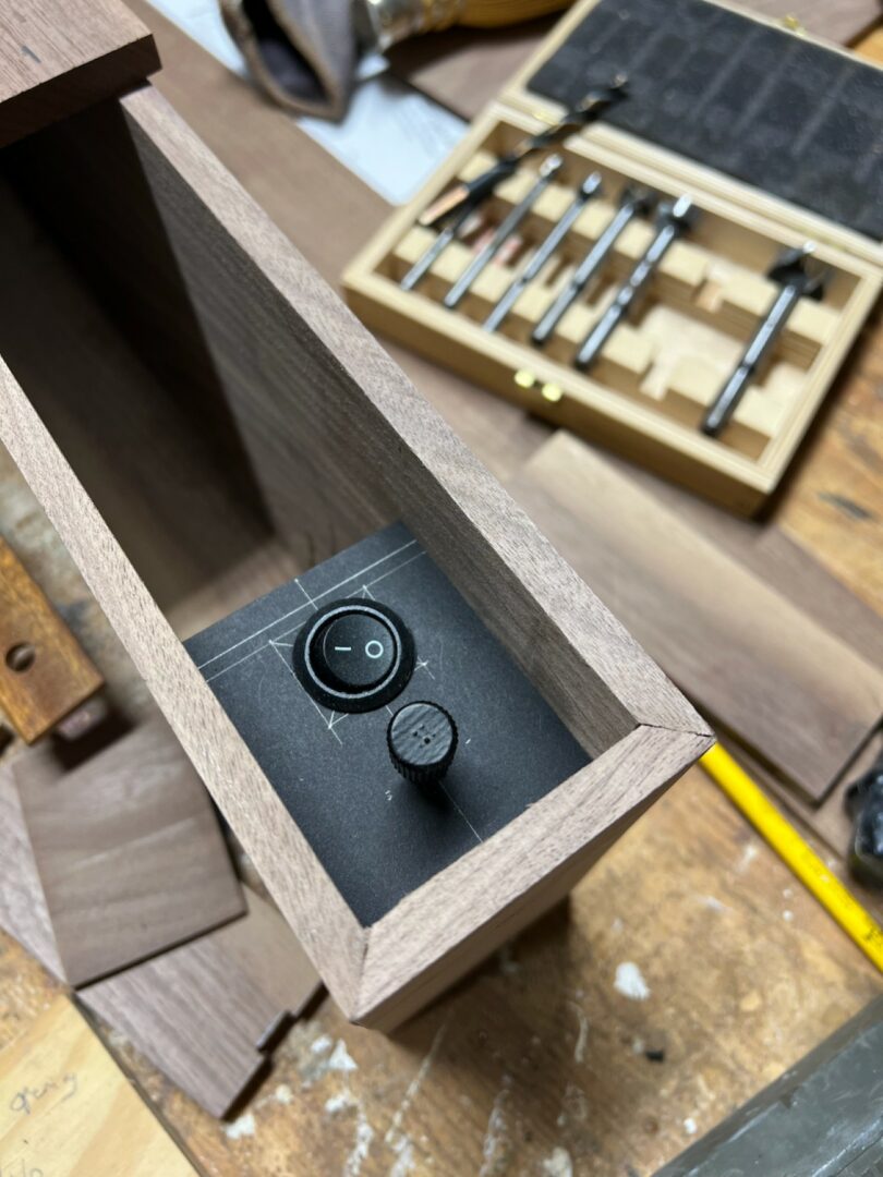 A close-up of a wooden box with a round on/off switch and a small knob mounted on a black panel; woodworking tools and materials are visible in the background