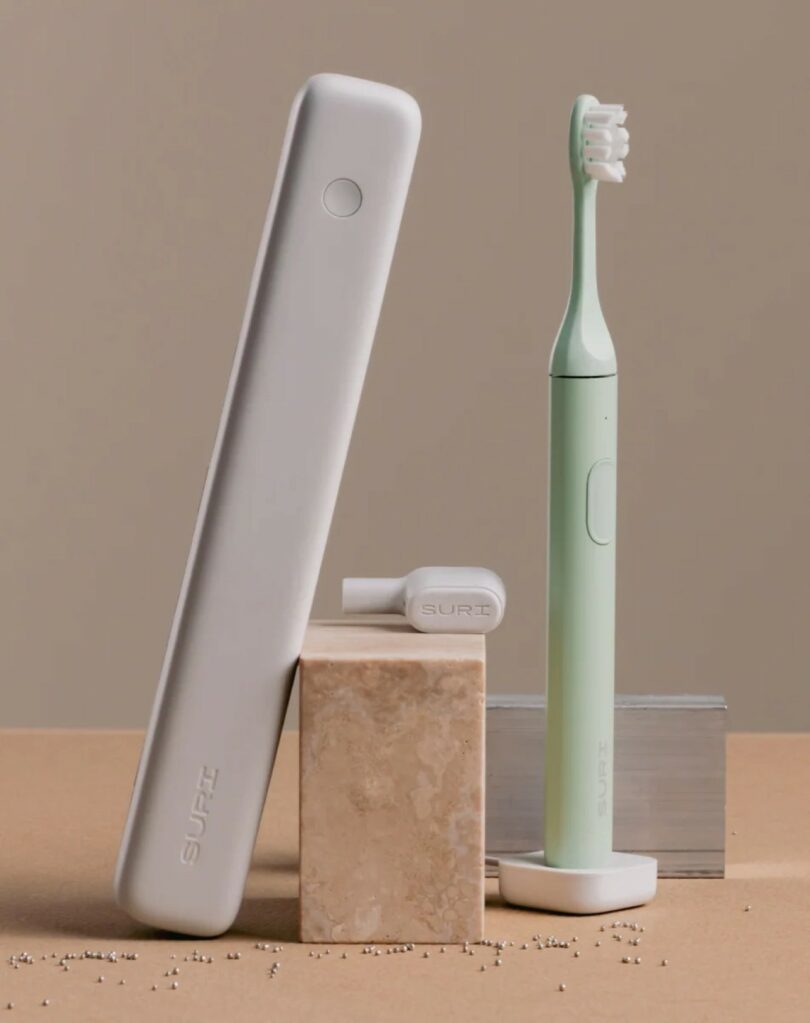 Un cepillo de dientes eléctrico de color verde menta con cabezal extraíble, un estuche de viaje blanco y una piedra rectangular están estratégicamente dispuestos sobre una superficie beige: una escena que sería perfecta para la guía de regalos Design Milk.