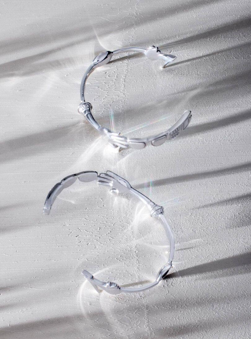 Dos pulseras de plata con extremos en forma de mano se colocan sobre una superficie blanca texturizada, y la luz crea suaves sombras y reflejos alrededor de las pulseras.