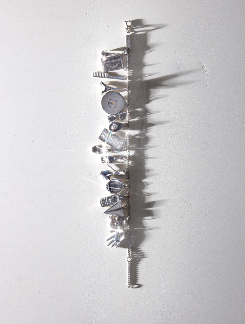 La pulsera de dijes de plata presenta una variedad de delicados dijes que incluyen una cámara, un corazón, un libro y una llave, colocados sobre una superficie gris claro con sombras visibles.