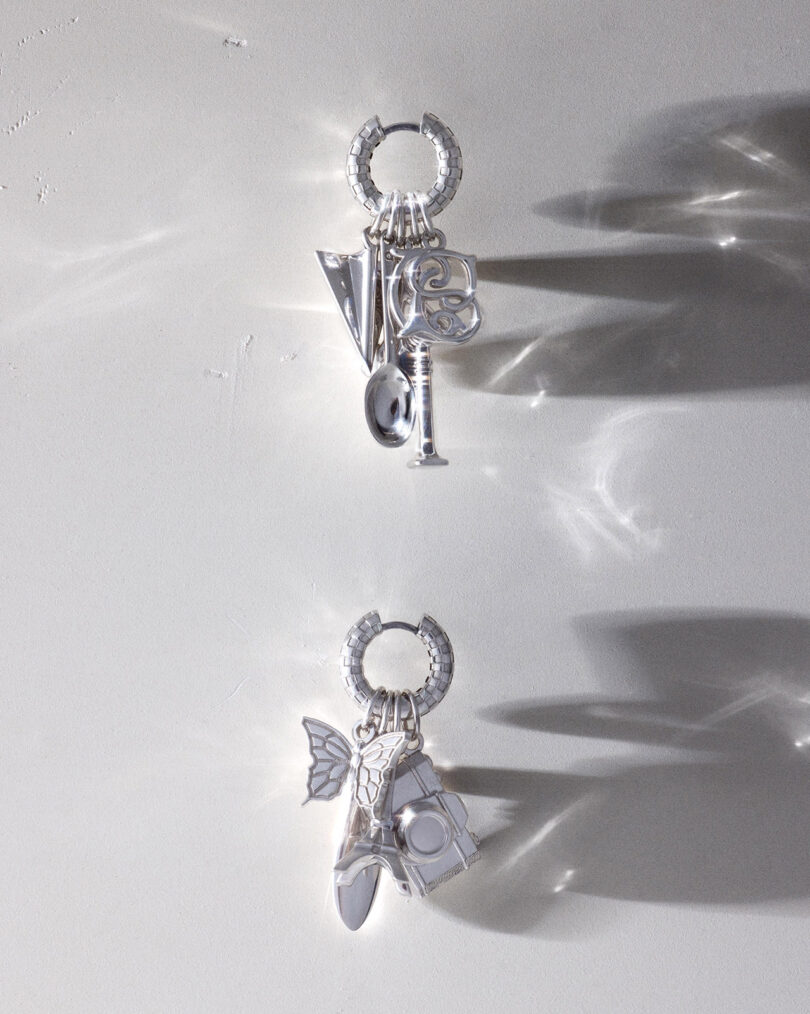 Dos aretes de aro plateados, cada uno con múltiples dijes de metal, se muestran sobre un fondo gris claro con sombras y reflejos.