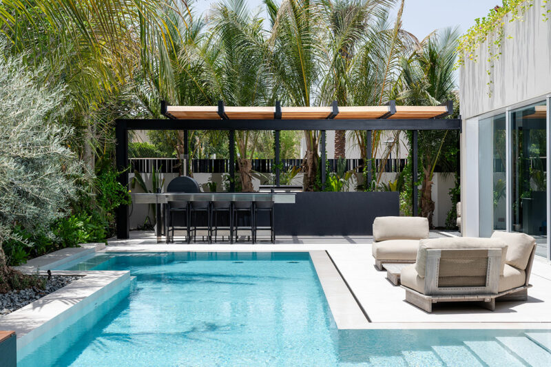 El moderno patio trasero cuenta con una piscina, tumbonas junto a la piscina y un área de cocina al aire libre debajo de una pérgola rodeada de palmeras.