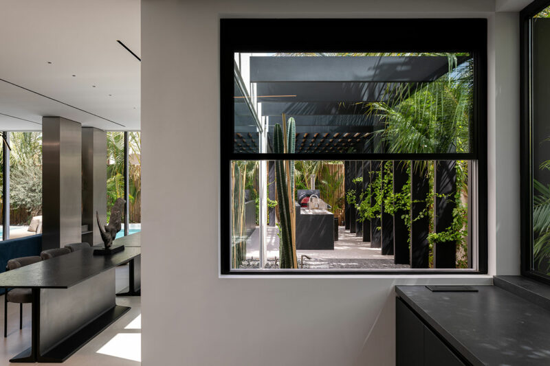 Interior moderno con grandes ventanales que dan a una terraza exterior con pérgola negra, exuberante vegetación y luz natural.