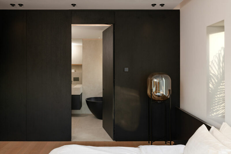 Dormitorio moderno con paredes oscuras y puerta que conduce a un baño minimalista con bañera negra; una lámpara de cristal se encuentra junto a la cama blanca.