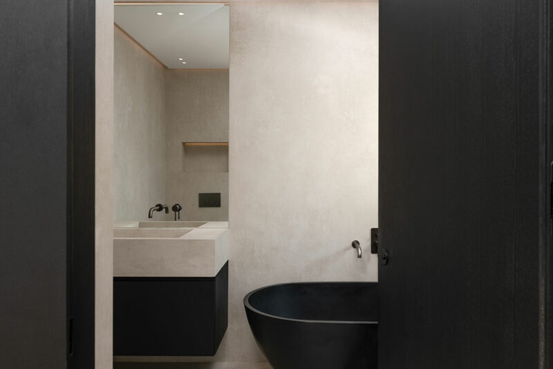 El baño minimalista presenta paredes beige, lavabo y encimera rectangulares, gabinetes negros, accesorios de pared negros y una bañera ovalada independiente negra.