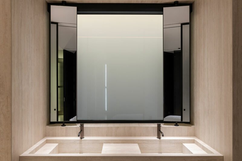 El tocador de baño con lavabo doble cuenta con dos grifos, un lavabo de piedra rectangular y un gran espejo moderno con panel deslizante en tonos neutros.