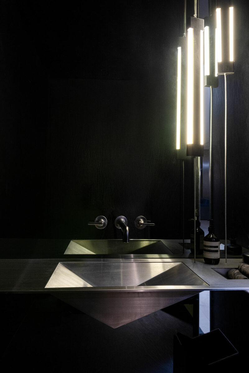 Cuarto de baño moderno con lavabo geométrico de metal, grifo de pared, paredes oscuras y luces de tubo verticales reflejadas en el espejo.