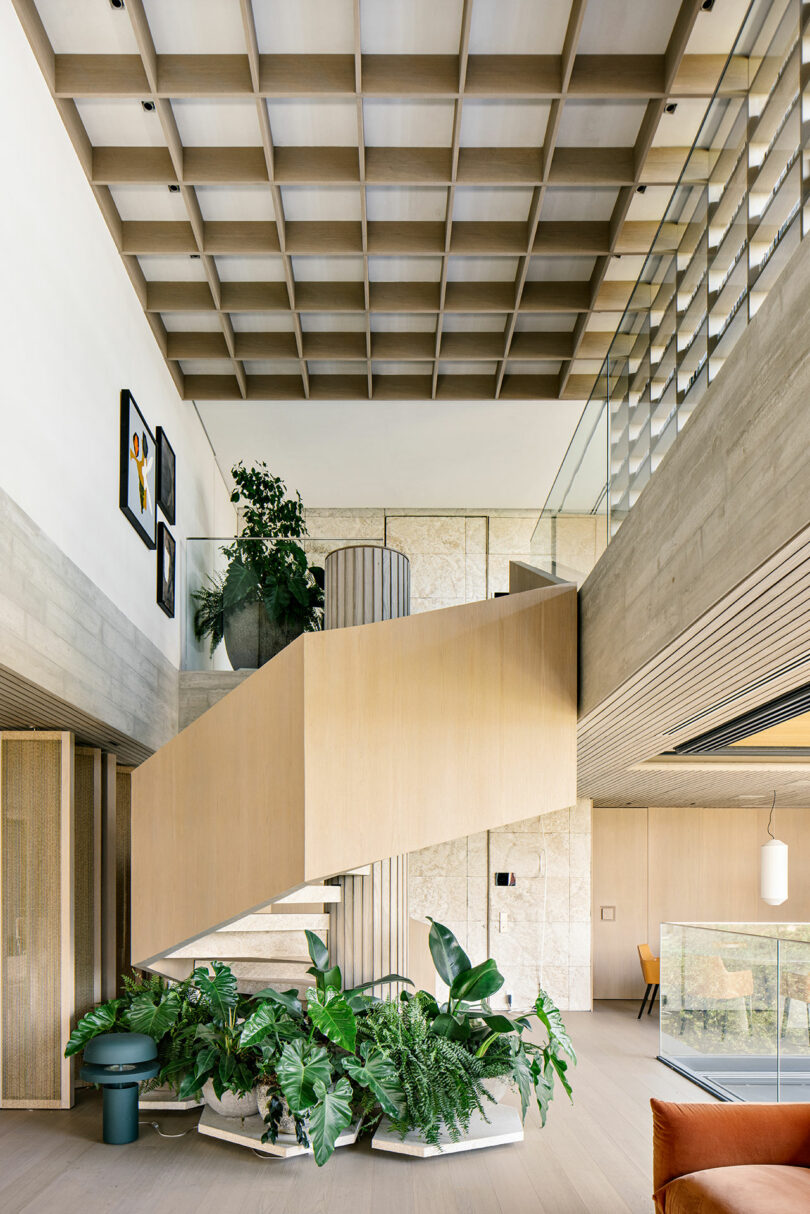 Interior moderno con escalera central de madera, grandes plantas en macetas y luz natural. Presenta techos de rejilla, barandillas de cristal y una decoración minimalista. Se pueden ver obras de arte en las paredes superiores.