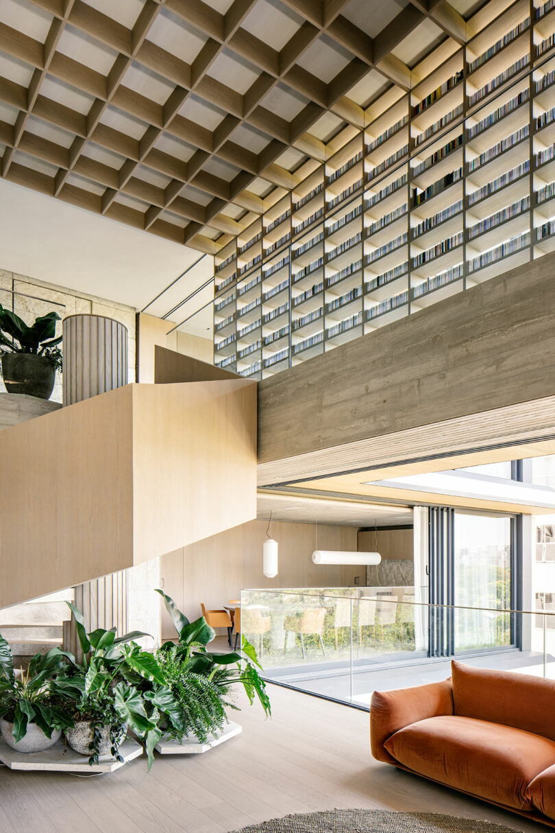 Interior moderno, sala de estar amplia y luminosa con techo geométrico, estantería grande, escalera abierta, plantas de interior y sofá naranja, con ventanales del suelo al techo.