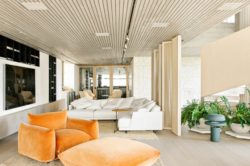 Sala de estar moderna con un gran sofá seccional blanco, sillas y reposapiés de terciopelo naranja, plantas de interior, estanterías empotradas y techo y suelo con paneles de madera.