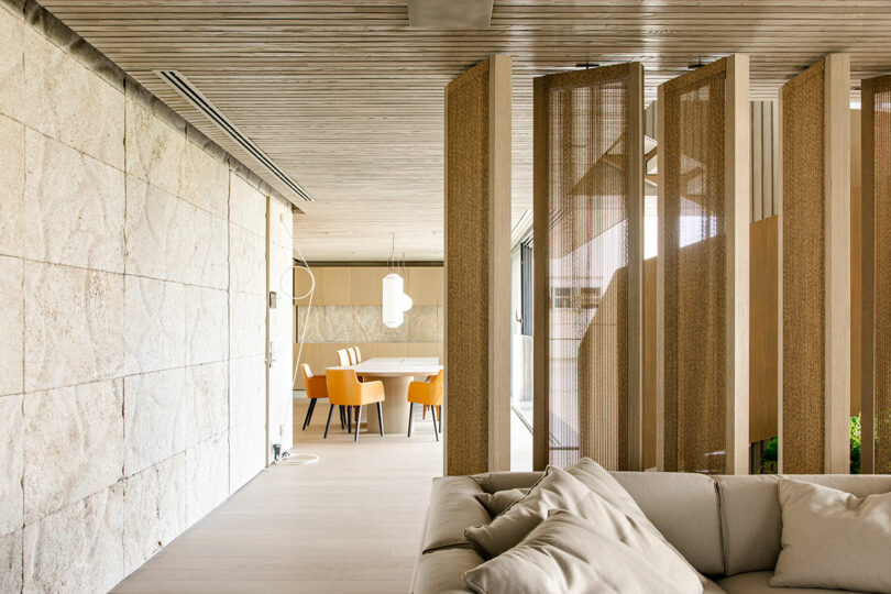 Sala de estar moderna con sofá beige, paredes de piedra texturizada, tabiques de madera y zona de comedor con sillas naranjas y una mesa de madera clara al fondo.
