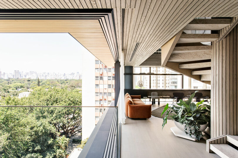 Interior moderno del apartamento con paneles de madera, escalera abierta, grandes ventanales, sofá marrón, plantas interiores y vistas a los árboles y edificios de la ciudad en el exterior.