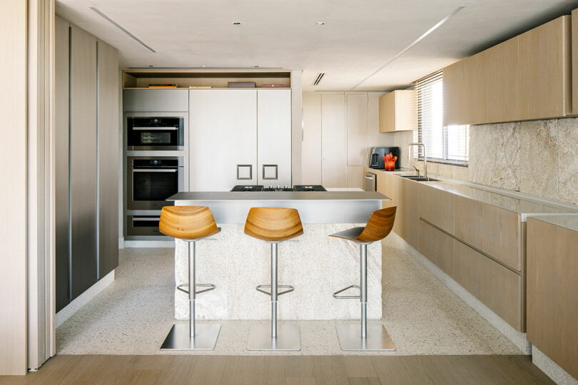 La moderna cocina cuenta con gabinetes de madera clara, una isla central con encimeras de mármol, tres taburetes de madera, un horno empotrado y grandes ventanales con contraventanas.