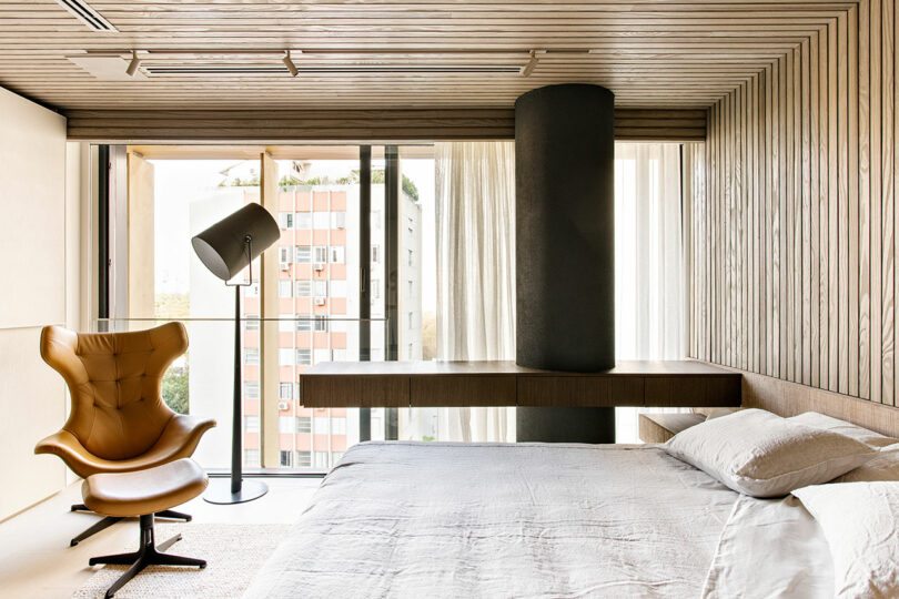 Dormitorio moderno con paredes y techo con paneles de madera, cama grande, sillón color canela, lámpara de pie y ventanales con vistas al edificio de apartamentos.
