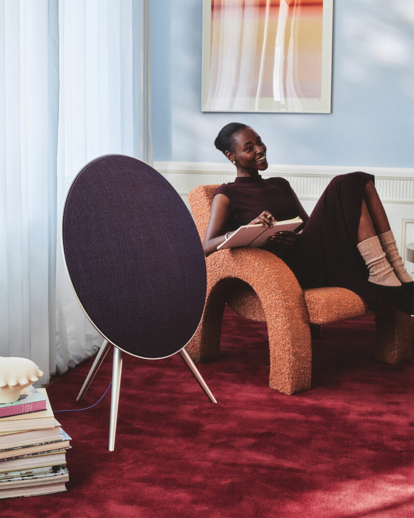 A woman sits on a textured chair in a room with red carpet, holding a tablet and pen beside a round speaker and a stack of Centennial Collection books.