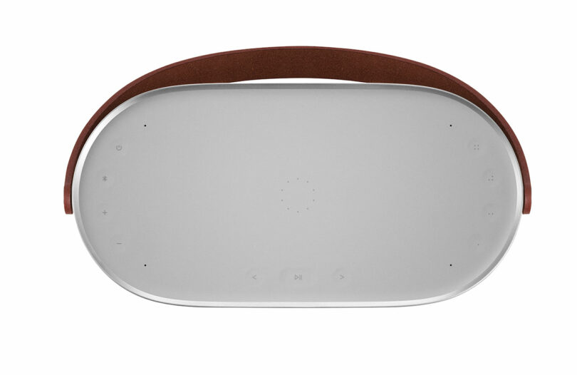 Oval-shaped wireless speaker with a brown handle, featuring touch-sensitive buttons for power, volume, and playback on the top surface.