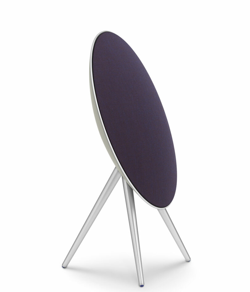 A modern speaker with a circular, dark fabric front and three silver legs in a tripod arrangement, set against a white background.