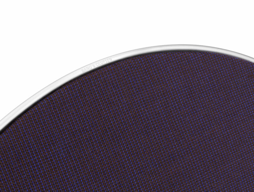 Close-up of the edge of a round speaker with a dark purple mesh cover and a silver rim, branded with "Bang & Olufsen.