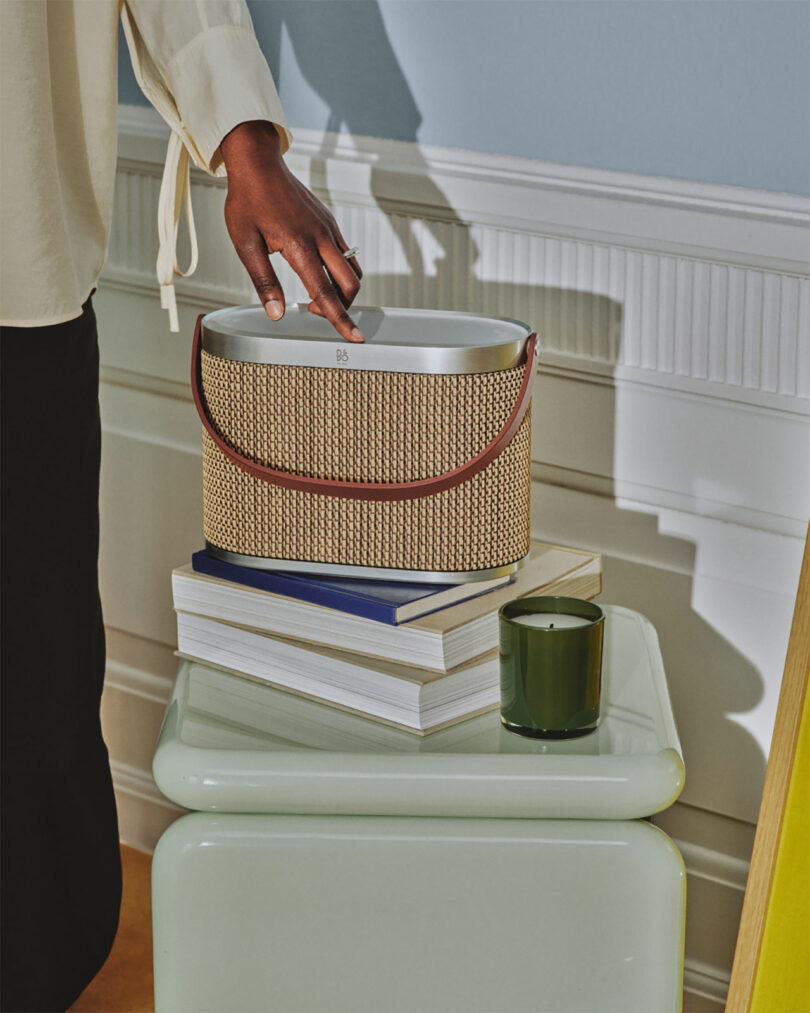 A person’s hand reaches into a woven handbag resting on a stack of books, next to a green candle on a light-colored table.