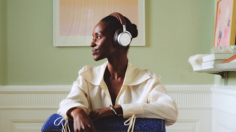 A person wearing over-ear headphones sits indoors, looking to the side, with a large blue bag on their lap and framed art on the wall behind them.