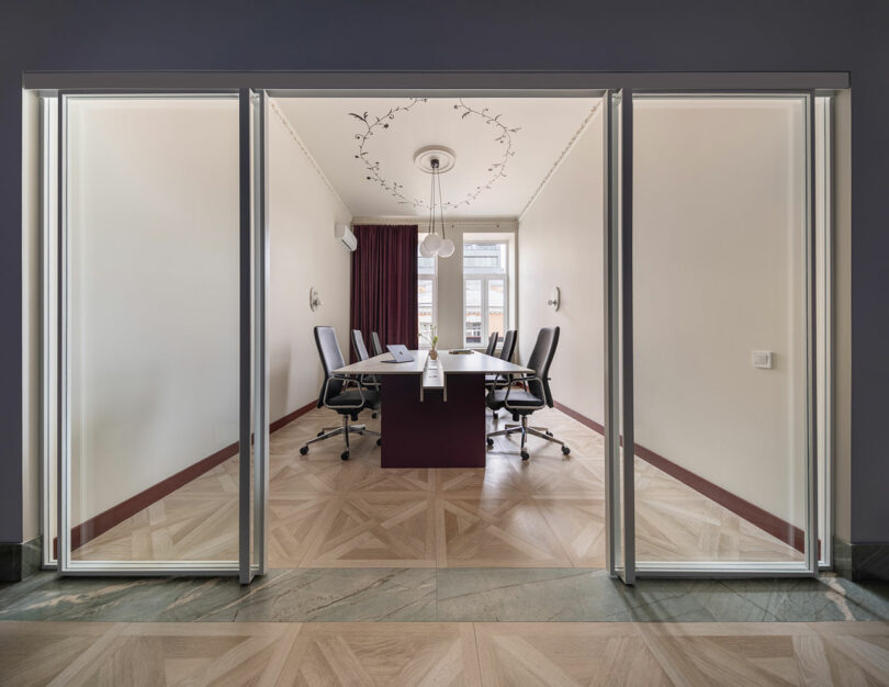 Moderna sala de conferencias con seis sillas de oficina alrededor de una mesa rectangular, grandes ventanales con cortinas, lámpara de araña y puertas correderas de cristal.
