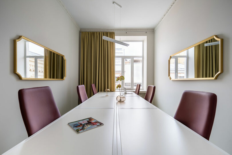 Una moderna sala de conferencias con sillas granates, una larga mesa blanca, dos espejos con marcos dorados, grandes ventanales con cortinas beige y una revista sobre la mesa.