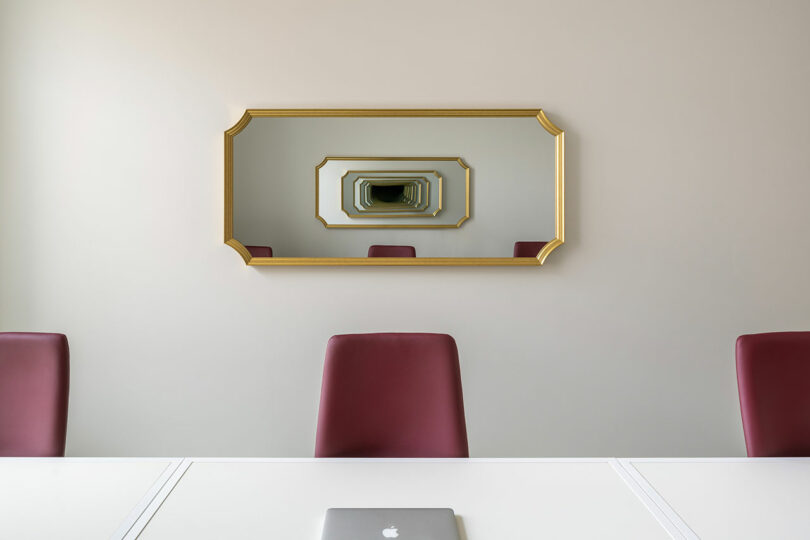 Una sala de conferencias con sillas rojas, mesa blanca, computadora portátil cerrada y un espejo con marco dorado que refleja la sala y a sí mismo varias veces.