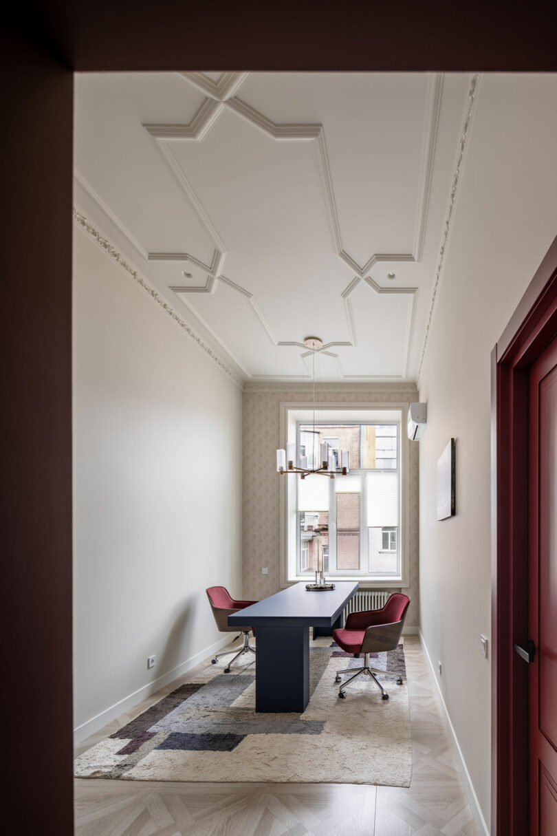 Una oficina moderna con un escritorio negro, dos sillas rojas, una alfombra estampada, una lámpara de araña y un gran ventanal que deja entrar la luz natural.