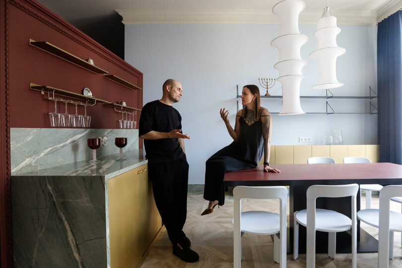Un hombre y una mujer conversan en una cocina moderna; la mujer está sentada en el mostrador y el hombre está de pie, uno frente al otro. La cocina está decorada en rojo, oro y mármol.