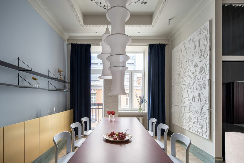 El comedor moderno cuenta con una larga mesa roja, ocho sillas plateadas, una lámpara de araña escultural blanca, arte en las paredes, estantes y una gran ventana con cortinas azules.
