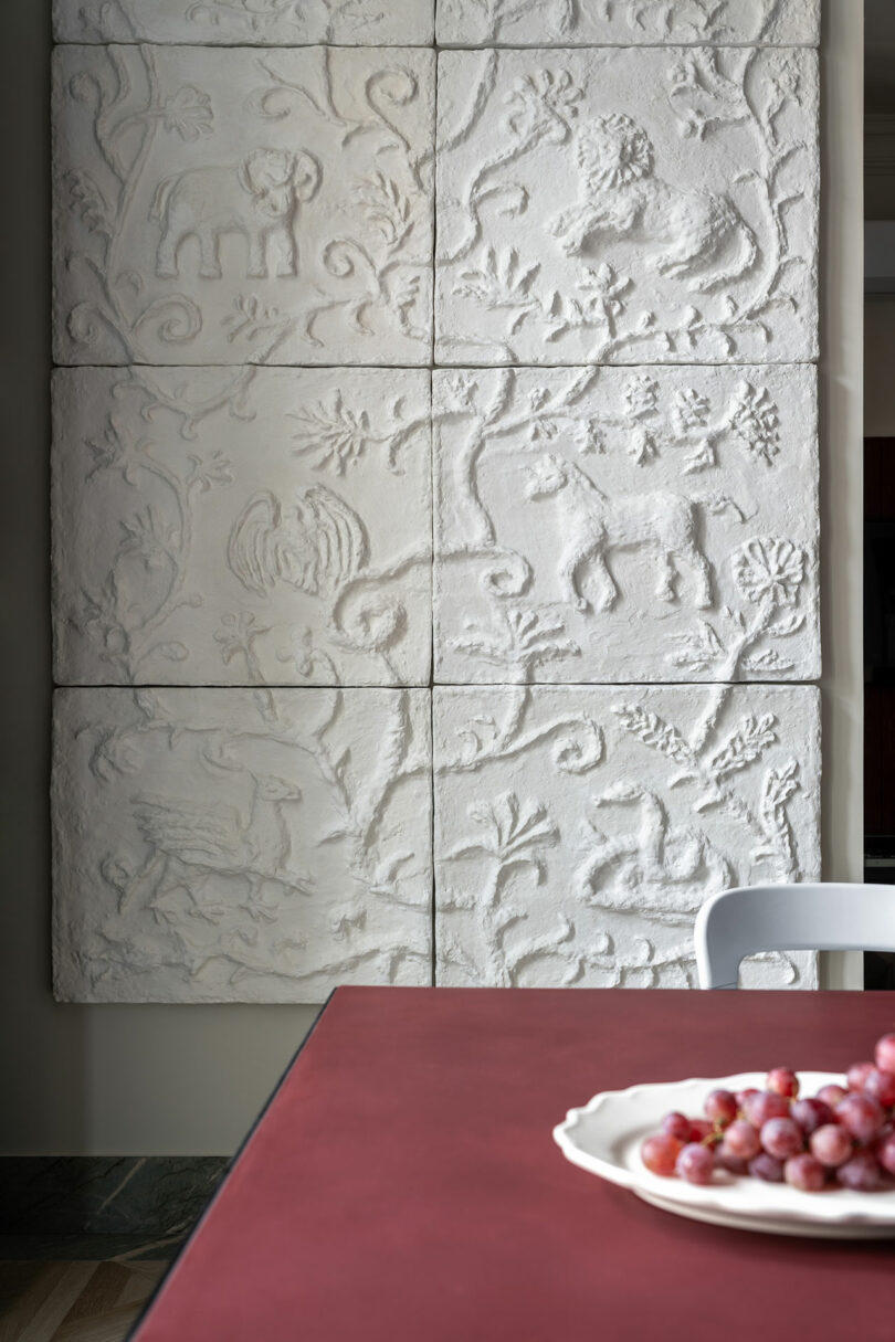 Detrás de la mesa color burdeos hay un panel de pared blanco texturizado con tallas de animales y hojas, con un plato de uvas blancas y una silla blanca en primer plano.