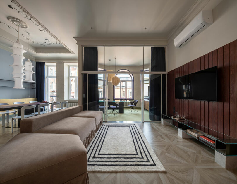 Sala de estar moderna con sofá seccional marrón, alfombra geométrica, TV montada en la pared, paneles de madera y una gran puerta de vidrio que conduce a un luminoso comedor.