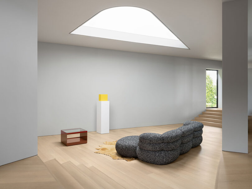 Salón minimalista con tragaluz geométrico, moderno sofá gris, mesa baja roja, pedestal con objetos amarillos, suelo de madera clara y paredes neutras. La luz natural llena todo el espacio.