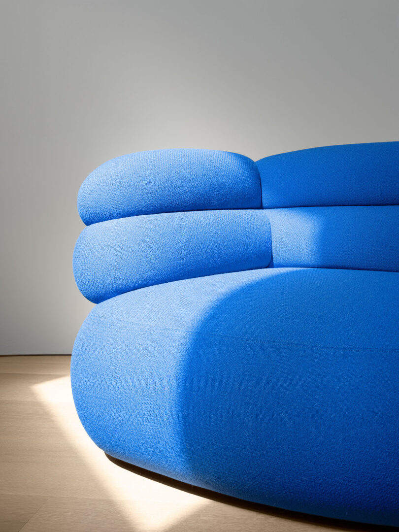 Un moderno sofá azul curvo con cojines en capas se asienta sobre un piso de madera clara contra una pared gris lisa iluminada por luz natural.