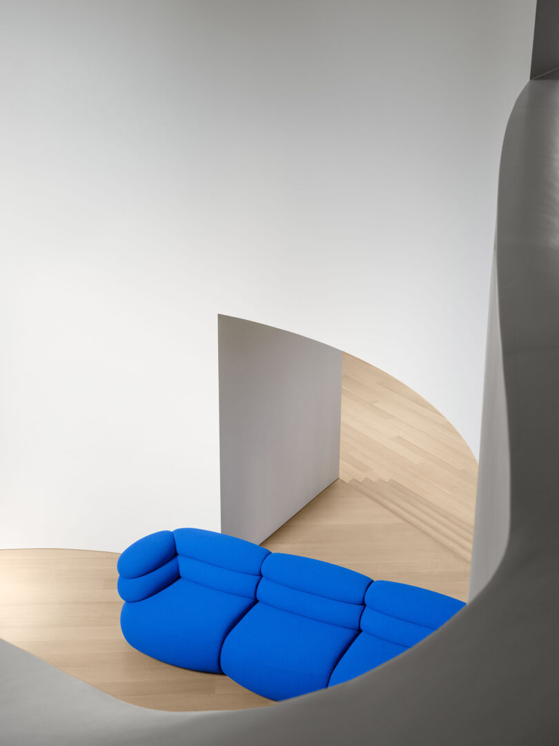Un sofá moderno de color azul brillante se asienta sobre un suelo de madera clara junto a una pared blanca curva y una puerta abierta para un interior minimalista.