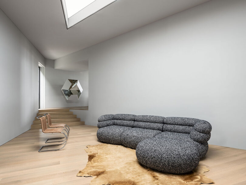 Salón minimalista con moderno sofá de textura gris, tres sillas beige, alfombra marrón, suelo de madera y arte geométrico en la pared bajo un tragaluz.