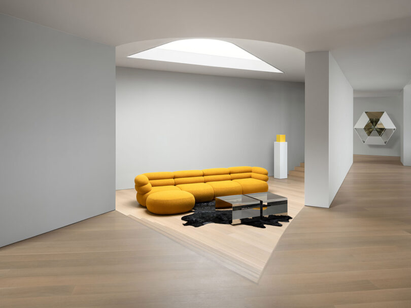 Salón moderno y minimalista con sofá amarillo, dos mesas cuadradas de cristal, moqueta negra y suelo de madera clara bajo un tragaluz geométrico.