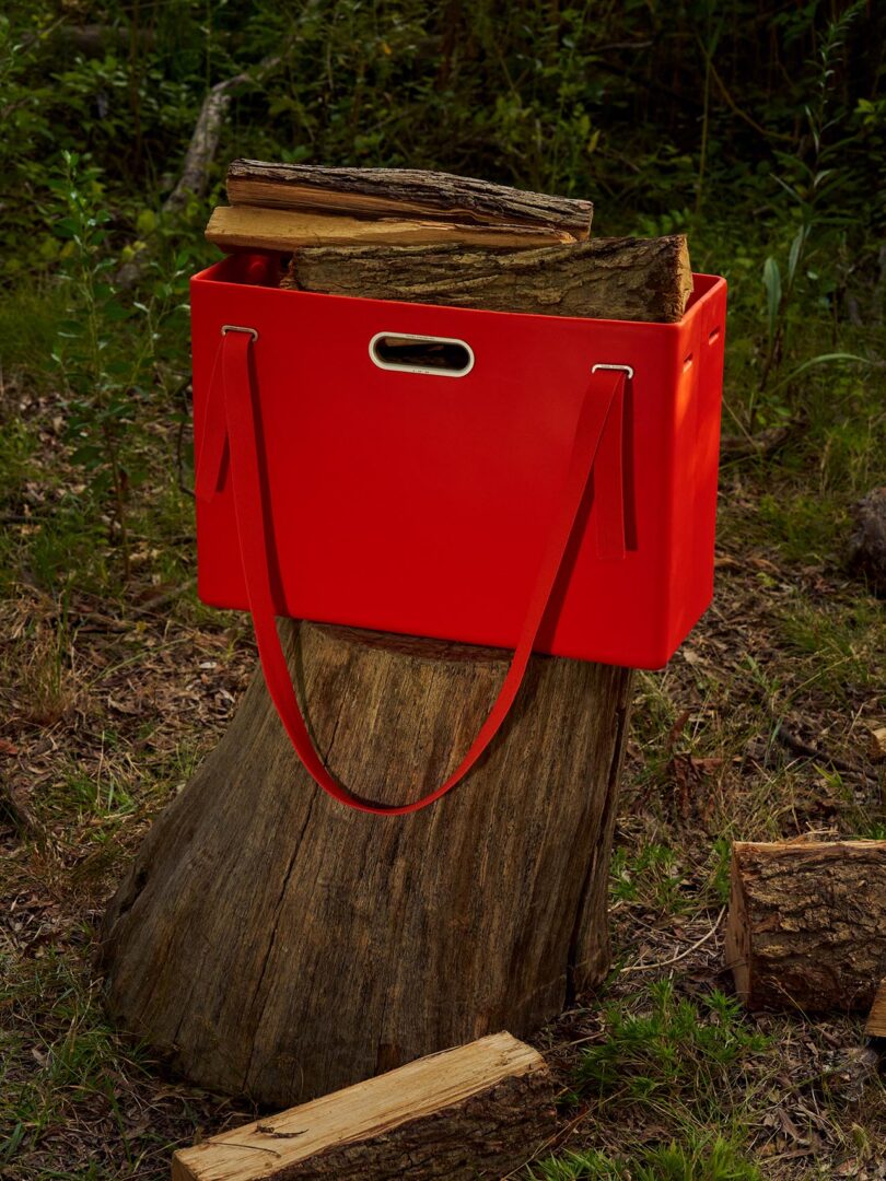 Una bolsa rectangular roja con asas llenas de leña se coloca sobre un tocón de árbol en el césped al aire libre.