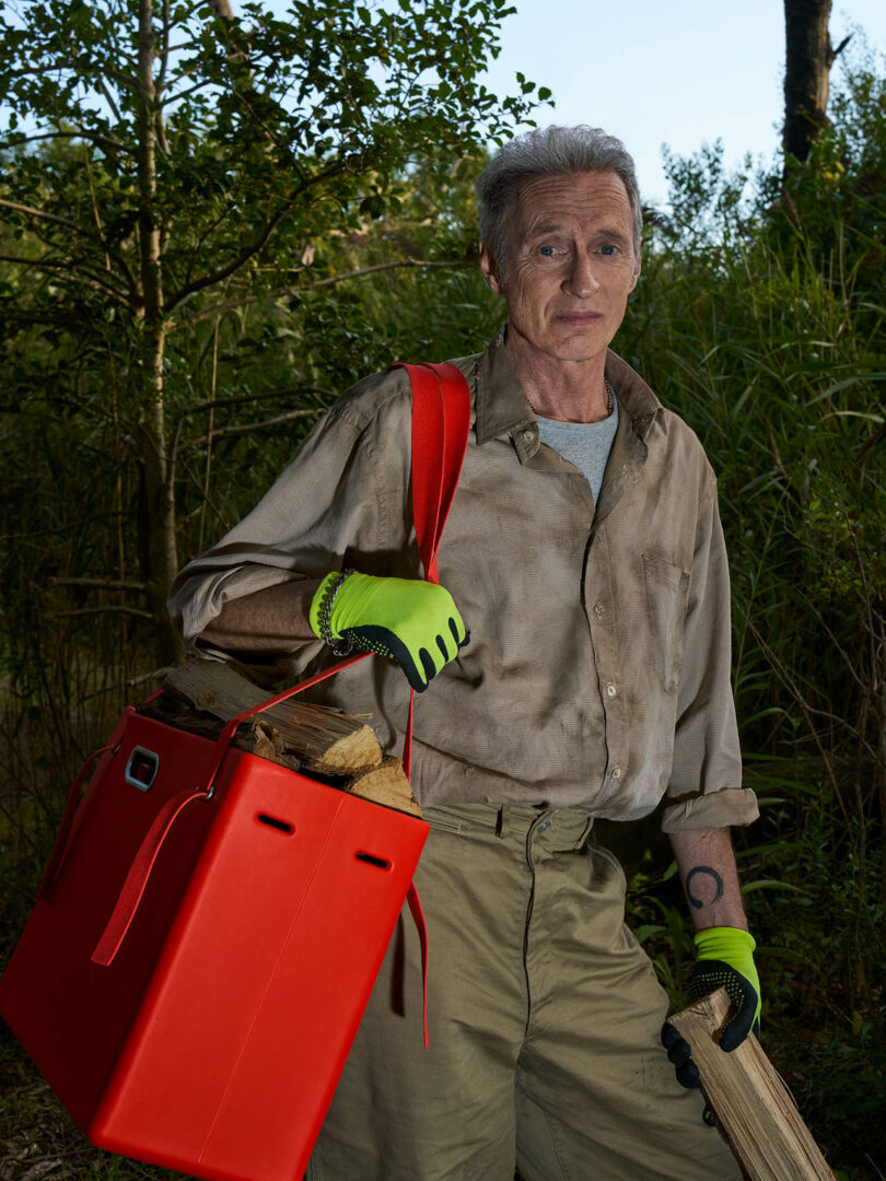 Un hombre vestido con una camisa sucia y guantes verdes lleva leña y un gran contenedor rojo en una zona boscosa al aire libre.