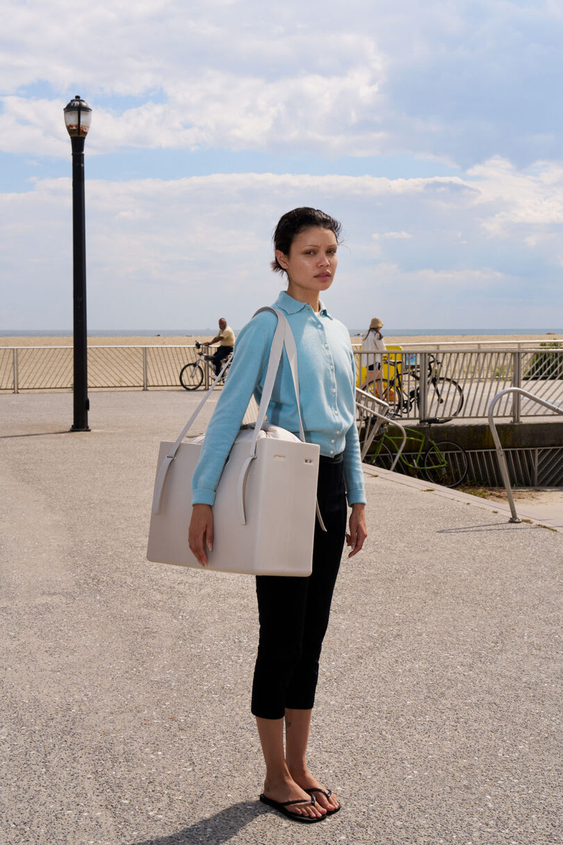 Un hombre vestido con una camisa azul claro, pantalones negros y chanclas está parado al aire libre con una gran bolsa blanca en la mano, con la playa y el paseo marítimo al fondo.