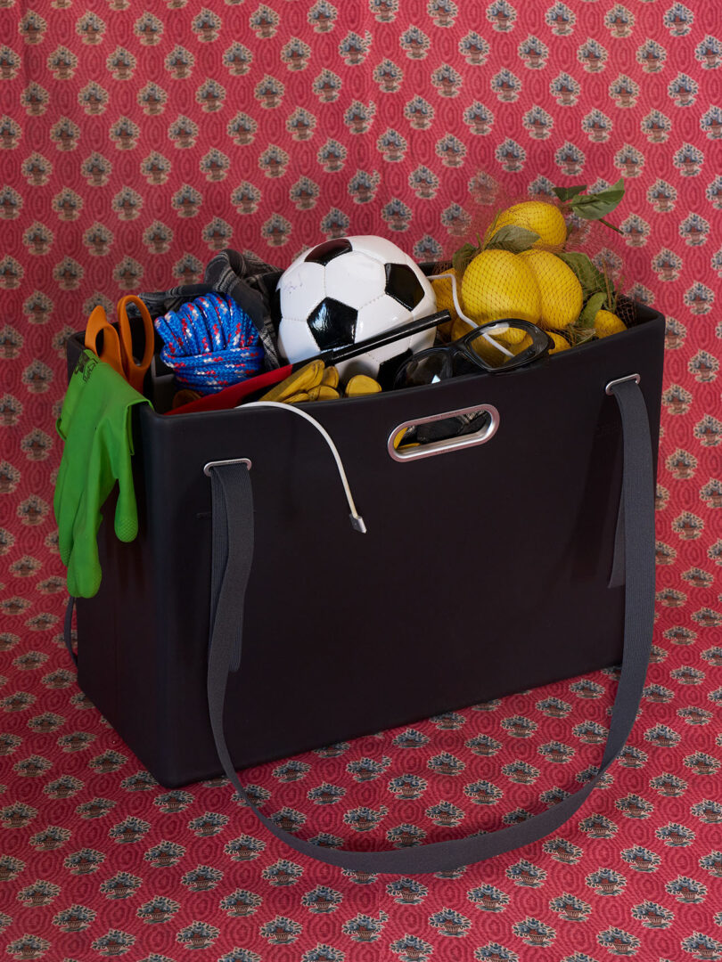 Frente a un fondo rojo estampado hay un sistema de bolsa PARSEL EVA negro lleno de balones de fútbol, ​​limones, guantes de jardinería, tijeras, cuerdas y otros artículos diversos.