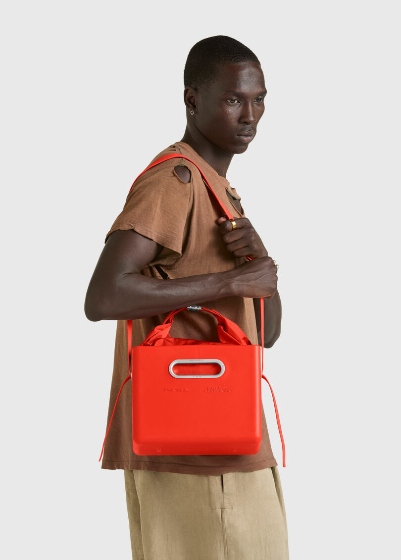 Un hombre vestido con una camisa marrón y pantalones beige sostiene un bolso tote PARSEL EVA Tote System de color rojo brillante con una correa para el hombro sobre un fondo claro.
