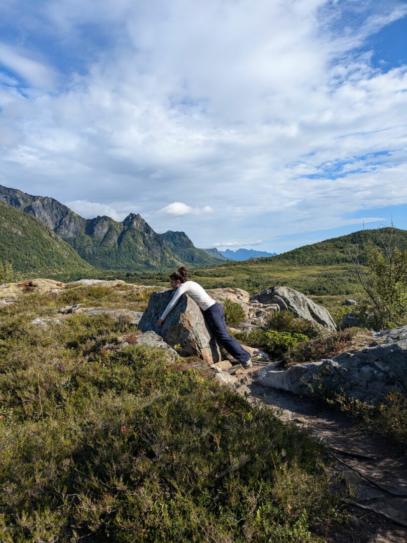 Bajo un cielo parcialmente nublado, una figura se inclina hacia adelante para abrazar una gran roca en un pintoresco paisaje montañoso, que recuerda las obras introspectivas de Dana Harel.