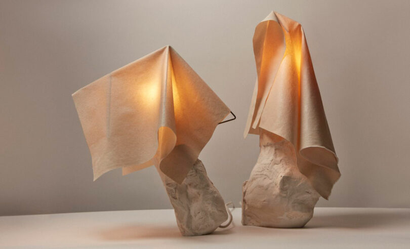 Dos lámparas de mesa esculturales de Dana Harel cuentan con bases blancas texturizadas y tela que cubre las fuentes de luz, proyectando un brillo suave y cálido sobre un fondo terroso.